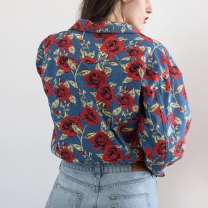 Pu&ograve; includere: Una camicia di jeans blu con un motivo floreale rosso e bianco. La camicia ha un colletto e maniche lunghe. La modella indossa la camicia con un paio di jeans blu chiaro.