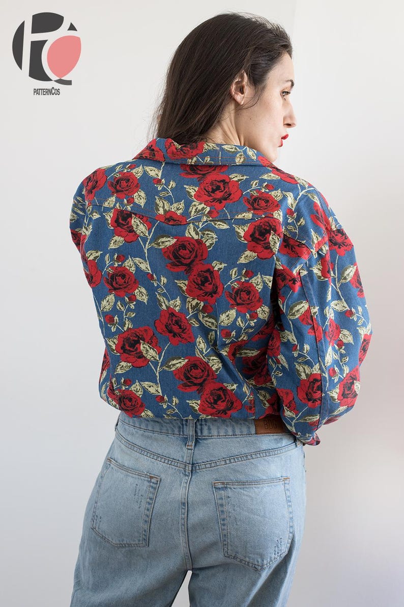 Pu&ograve; includere: Una camicia di jeans blu con un motivo floreale rosso e bianco. La camicia ha un colletto e maniche lunghe. La modella indossa la camicia con un paio di jeans blu chiaro.