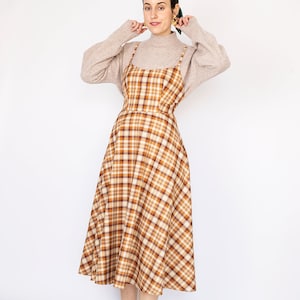 Pode incluir: Uma mulher vestindo um vestido midi xadrez marrom e bege com al&ccedil;as finas. Ela est&aacute; vestindo um su&eacute;ter de gola alta marrom claro sobre o vestido e t&ecirc;nis brancos. O vestido tem um corpete justo e uma saia levemente rodada.