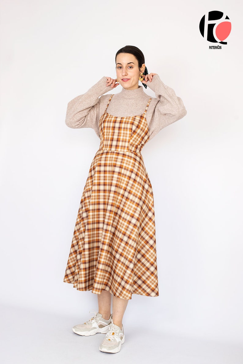 Pode incluir: Uma mulher vestindo um vestido midi xadrez marrom e bege com al&ccedil;as finas. Ela est&aacute; vestindo um su&eacute;ter de gola alta marrom claro sobre o vestido e t&ecirc;nis brancos. O vestido tem um corpete justo e uma saia levemente rodada.