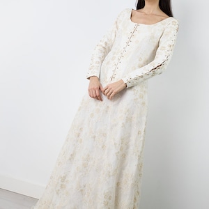 Peut inclure: Une robe maxi blanche avec une superposition de dentelle et des broderies dor&eacute;es. La robe a un col rond, des manches longues et un corsage ajust&eacute;. Les manches ont des d&eacute;tails en dentelle aux poignets. La robe a une jupe ample avec une frange sur l'ourlet.