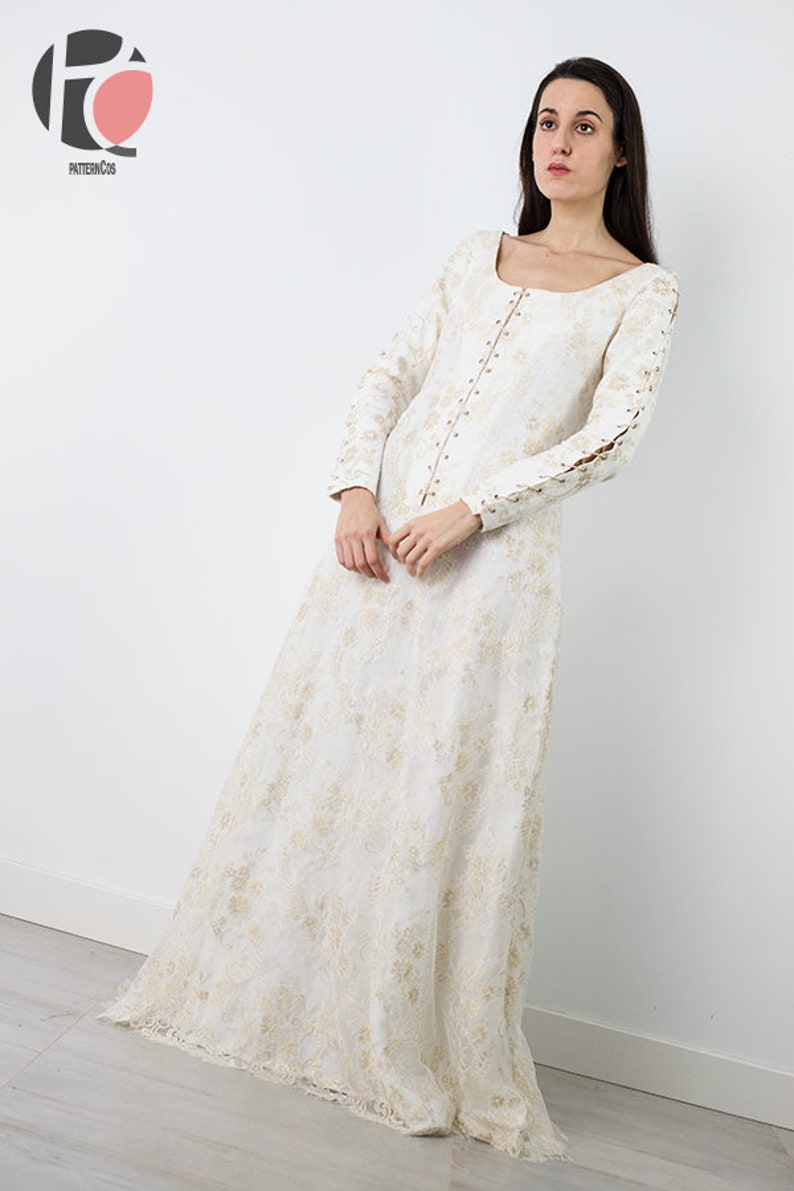 Peut inclure: Une robe maxi blanche avec une superposition de dentelle et des broderies dor&eacute;es. La robe a un col rond, des manches longues et un corsage ajust&eacute;. Les manches ont des d&eacute;tails en dentelle aux poignets. La robe a une jupe ample avec une frange sur l'ourlet.