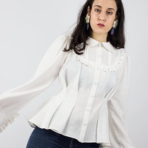 May include: A white button-up blouse with a ruffled collar and cuffs. The blouse has a fitted bodice and a flared skirt. The model is wearing blue jeans.