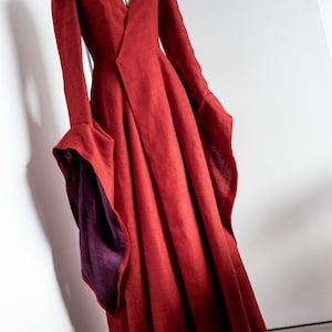 May include: A long, flowing red dress with a V-neck and long sleeves. The sleeves have a flared design with a contrasting purple lining. The dress is displayed on a mannequin against a white background, showcasing its elegant silhouette.