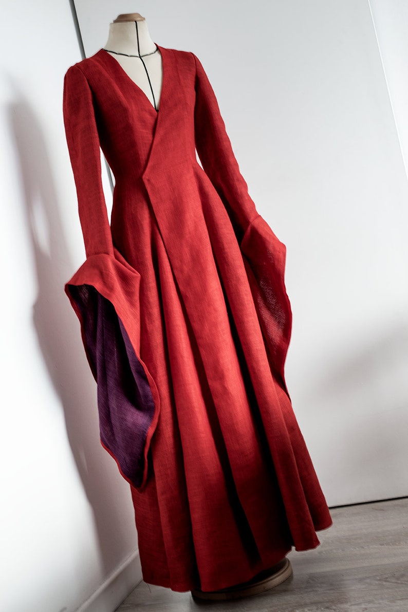 May include: A long, flowing red dress with a V-neck and long sleeves. The sleeves have a flared design with a contrasting purple lining. The dress is displayed on a mannequin against a white background, showcasing its elegant silhouette.