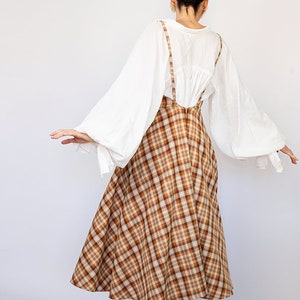 Pode incluir: Uma saia xadrez marrom e bege com uma blusa branca e um par de sand&aacute;lias verdes. A saia tem uma forma ampla e em A e &eacute; usada com uma blusa branca de mangas compridas e bufantes. As sand&aacute;lias s&atilde;o simples e t&ecirc;m um salto baixo.