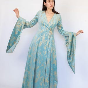 May include: A long, flowing dress in a light blue hue with a gold pattern. The dress features long, flared sleeves and a V-neckline. A gold-colored tie cinches the waist. The dress is made of a soft, velvety fabric, perfect for a special occasion.