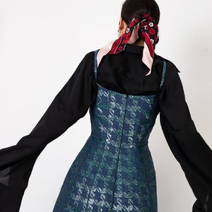 K&ouml;nnte beinhalten: Ein &auml;rmelloses, knielanges Kleid mit blauem und gr&uuml;nem Hahnentrittmuster. Das Kleid wird &uuml;ber einem schwarzen Langarmshirt getragen. Ein floraler Schal ist im Haar gebunden. Das Kleid hat ein figurbetontes Oberteil und einen ausgestellten Rock.