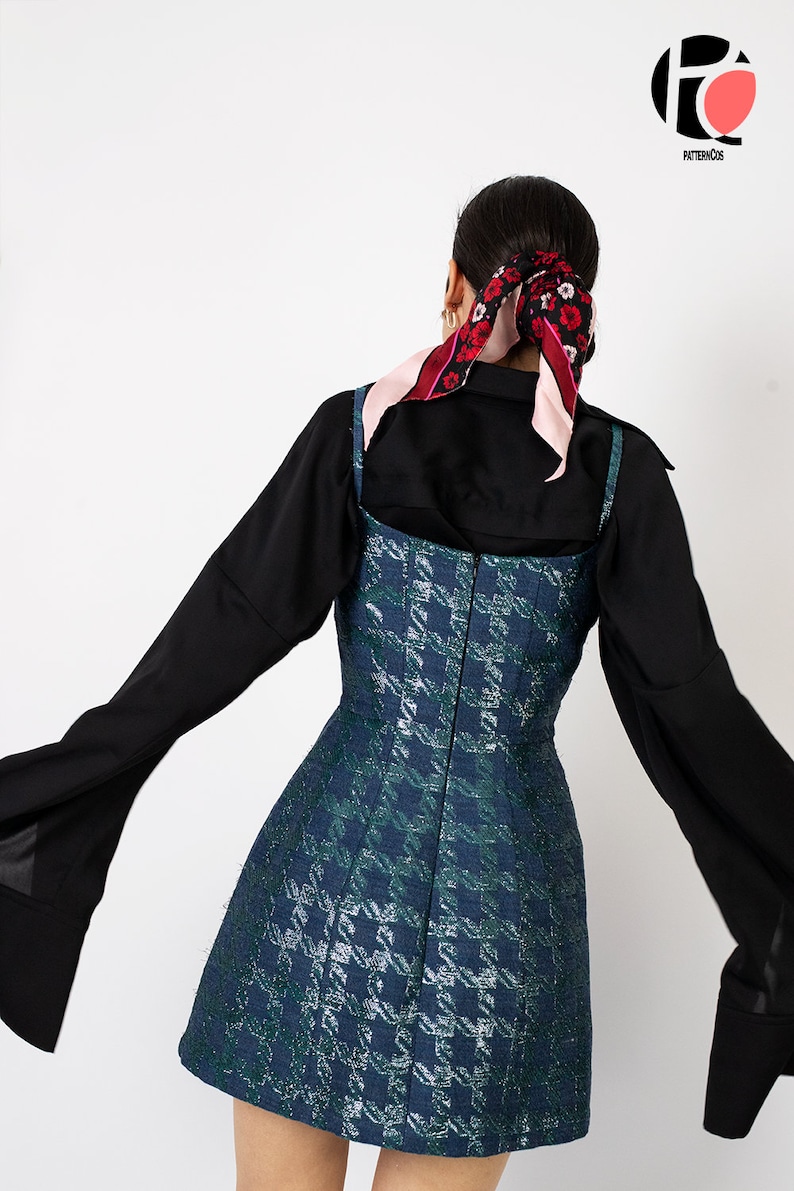 K&ouml;nnte beinhalten: Ein &auml;rmelloses, knielanges Kleid mit blauem und gr&uuml;nem Hahnentrittmuster. Das Kleid wird &uuml;ber einem schwarzen Langarmshirt getragen. Ein floraler Schal ist im Haar gebunden. Das Kleid hat ein figurbetontes Oberteil und einen ausgestellten Rock.