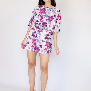 May include: A woman wearing a white and purple floral print top and matching shorts. She is wearing brown sandals and standing on a white background.