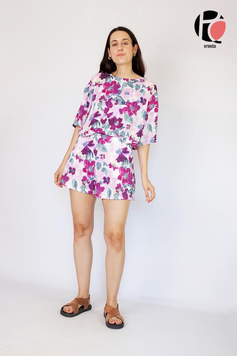 May include: A woman wearing a white and purple floral print top and matching shorts. She is wearing brown sandals and standing on a white background.