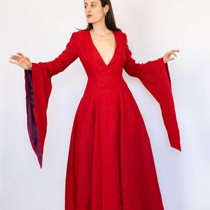 May include: A long, flowing red dress with a deep V-neck and dramatic bell sleeves. The sleeves have a purple lining. The dress is floor-length and made of a textured fabric. The dress is a bold statement piece.