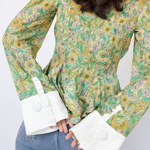 May include: A yellow and green floral print blouse with long sleeves and white ruffled cuffs. The cuffs have a button closure.