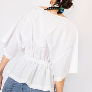 May include: A white, loose-fitting blouse with a gathered waist and short sleeves. The blouse is worn with blue jeans.