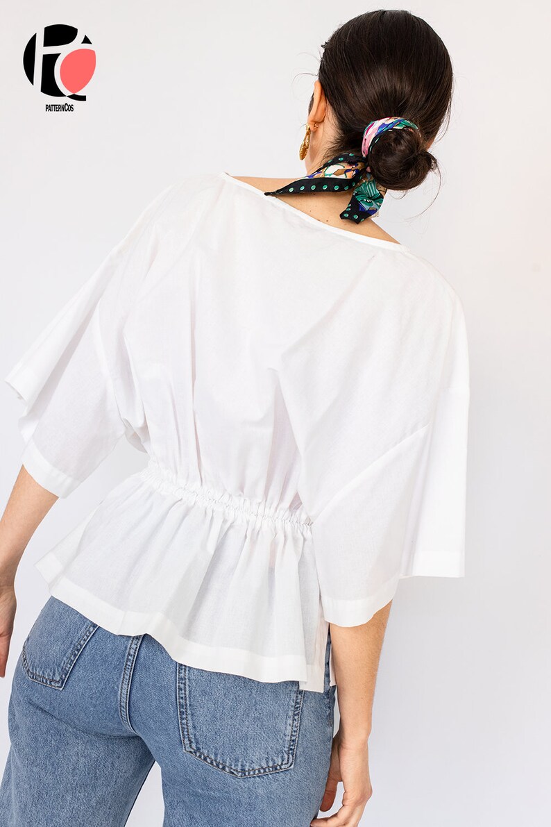 May include: A white, loose-fitting blouse with a gathered waist and short sleeves. The blouse is worn with blue jeans.