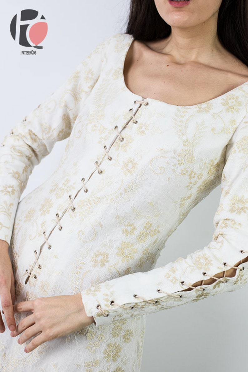 Peut inclure: Une robe blanche &agrave; motif de dentelle et accents dor&eacute;s. La robe a un devant &agrave; lacets et des poignets &agrave; lacets.