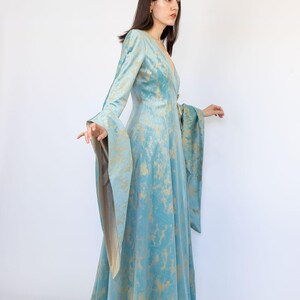 May include: A long, flowing teal dress with long, bell sleeves and a V-neck. The dress features a gold, abstract pattern throughout. The model is standing in front of a white background. The dress is a unique piece of apparel.