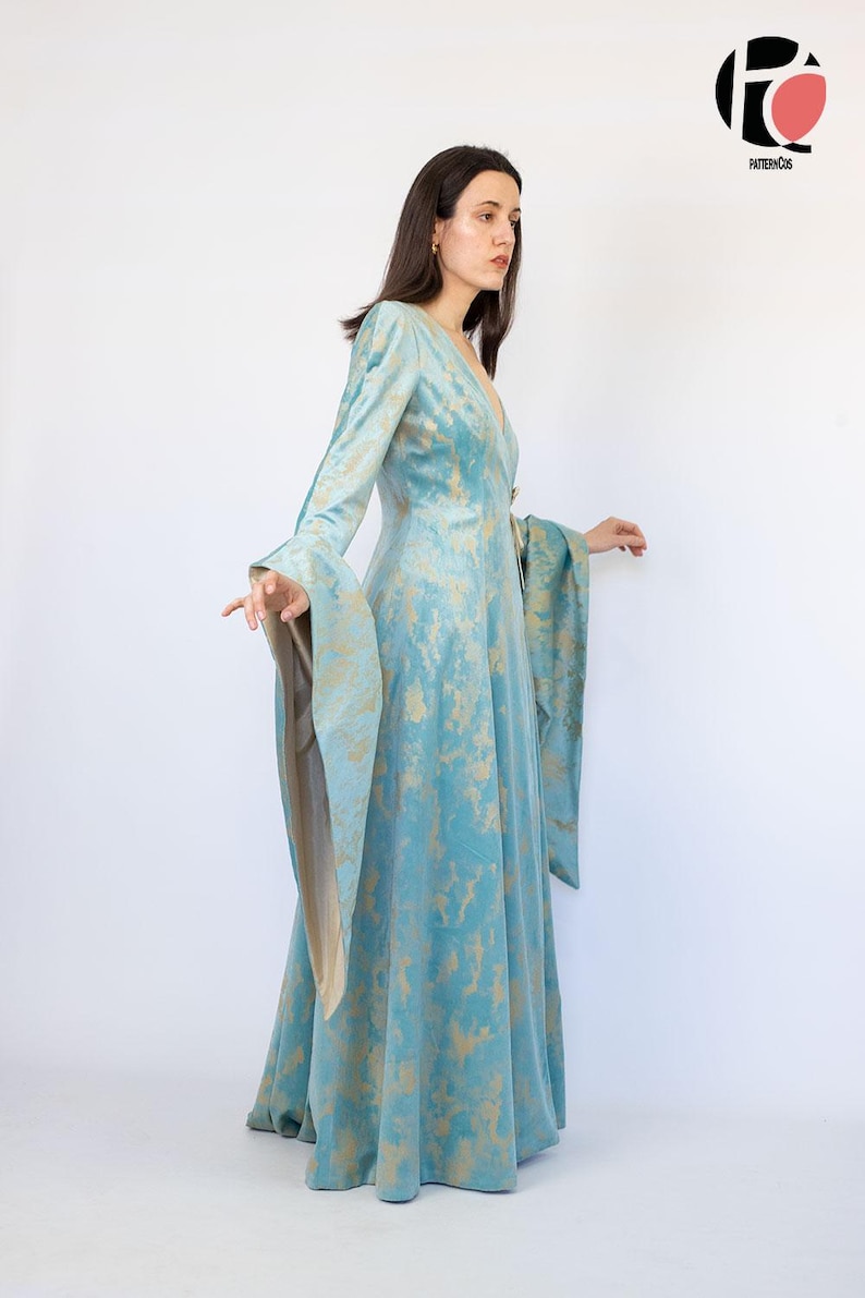 May include: A long, flowing teal dress with long, bell sleeves and a V-neck. The dress features a gold, abstract pattern throughout. The model is standing in front of a white background. The dress is a unique piece of apparel.