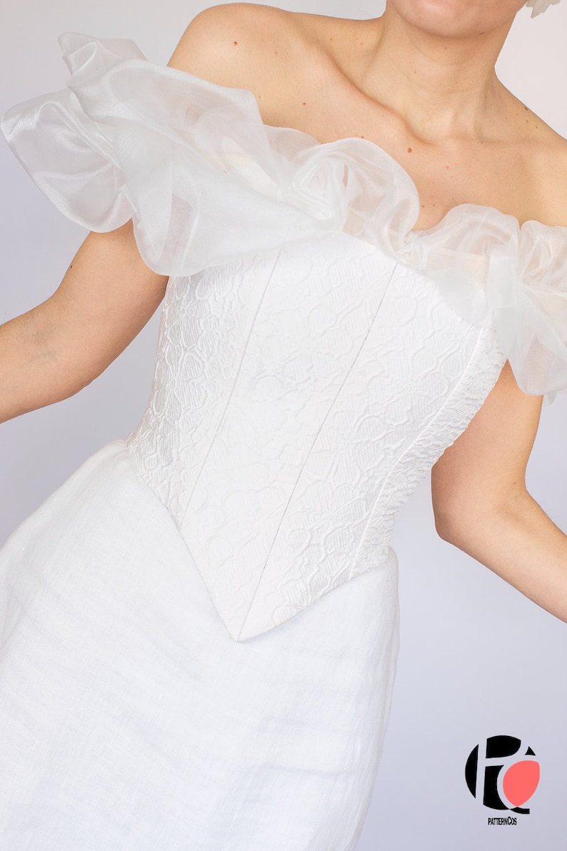 May include: A white off-the-shoulder wedding dress with a ruffled bodice and a fitted, lace-like corset. The skirt is flowing and has a simple, elegant design.