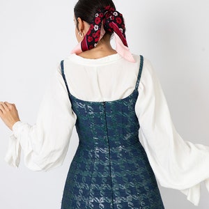 K&ouml;nnte beinhalten: Eine Frau tr&auml;gt eine wei&szlig;e Bluse mit langen &Auml;rmeln und ein dunkelgr&uuml;nes und blau gemustertes Kleid. Das Kleid hat d&uuml;nne Tr&auml;ger und ein eng anliegendes Oberteil. Ein floraler Schal ist im Haar der Frau gebunden.