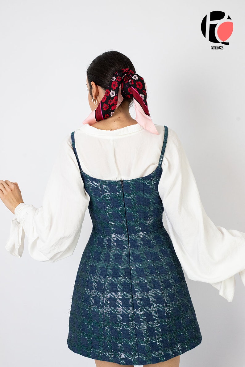 K&ouml;nnte beinhalten: Eine Frau tr&auml;gt eine wei&szlig;e Bluse mit langen &Auml;rmeln und ein dunkelgr&uuml;nes und blau gemustertes Kleid. Das Kleid hat d&uuml;nne Tr&auml;ger und ein eng anliegendes Oberteil. Ein floraler Schal ist im Haar der Frau gebunden.