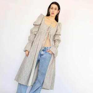 May include: A long, open-front dress with a floral pattern, featuring a square neckline and puffed sleeves. The dress is paired with light blue jeans. The image also includes a logo in the upper right corner.