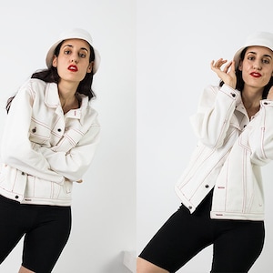 Pu&ograve; includere: Una donna indossa una giacca di jeans bianca con cuciture rosse, pantaloncini da ciclista neri e un cappello a secchiello bianco. Sta posando con le mani sulla testa.