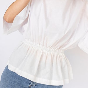 May include: A white, short-sleeved blouse with a gathered elastic waistband. The blouse is made of a lightweight fabric and has a relaxed fit.
