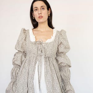 May include: A long-sleeved dress with a floral pattern in shades of beige and brown. The dress features a square neckline with white lace trim, a tie-up front, and voluminous sleeves. The dress is layered over a white garment.