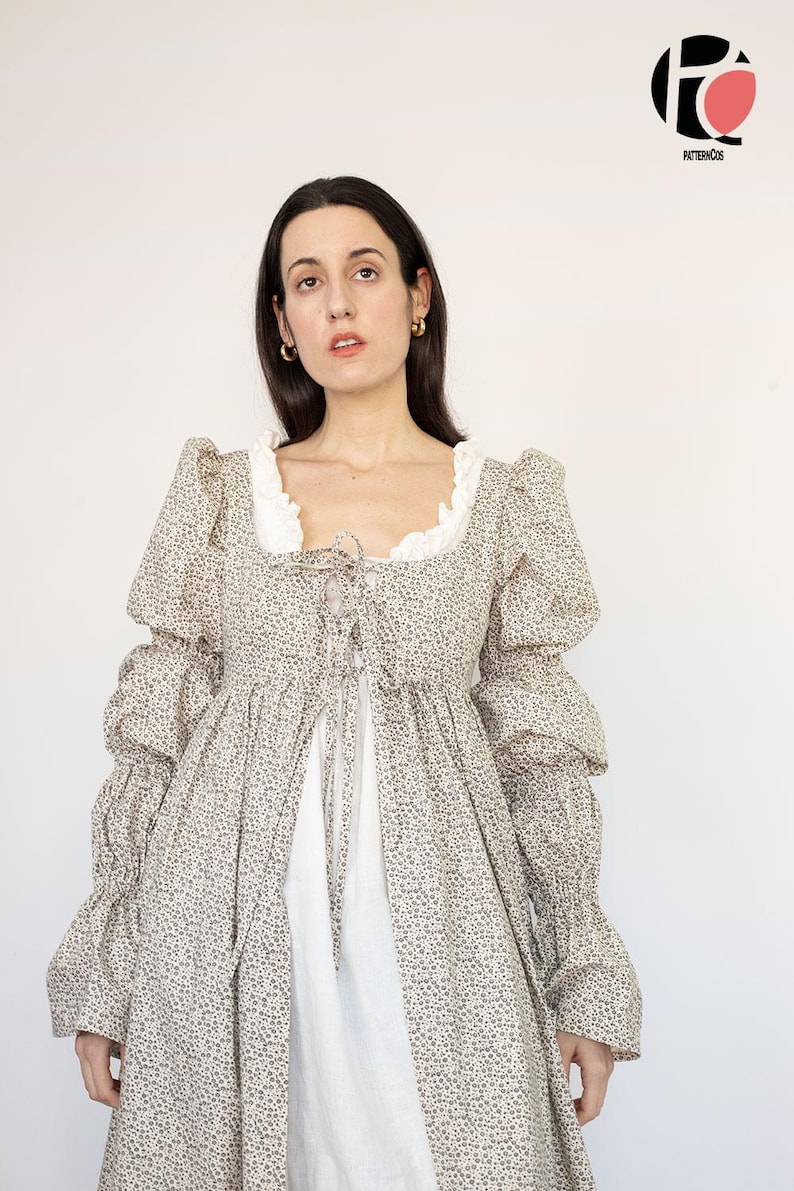 May include: A long-sleeved dress with a floral pattern in shades of beige and brown. The dress features a square neckline with white lace trim, a tie-up front, and voluminous sleeves. The dress is layered over a white garment.