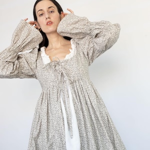 May include: A long-sleeved dress with a floral pattern in shades of gray and white. The dress features a square neckline with a white lace trim and a tie-up front. The sleeves are long and voluminous. The dress is a vintage style.