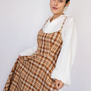 Pode incluir: Uma mulher vestindo um vestido de avental xadrez marrom e branco sobre uma camisa branca abotoada. O vestido tem um decote redondo e al&ccedil;as finas. A mulher est&aacute; segurando a saia do vestido com a m&atilde;o direita.