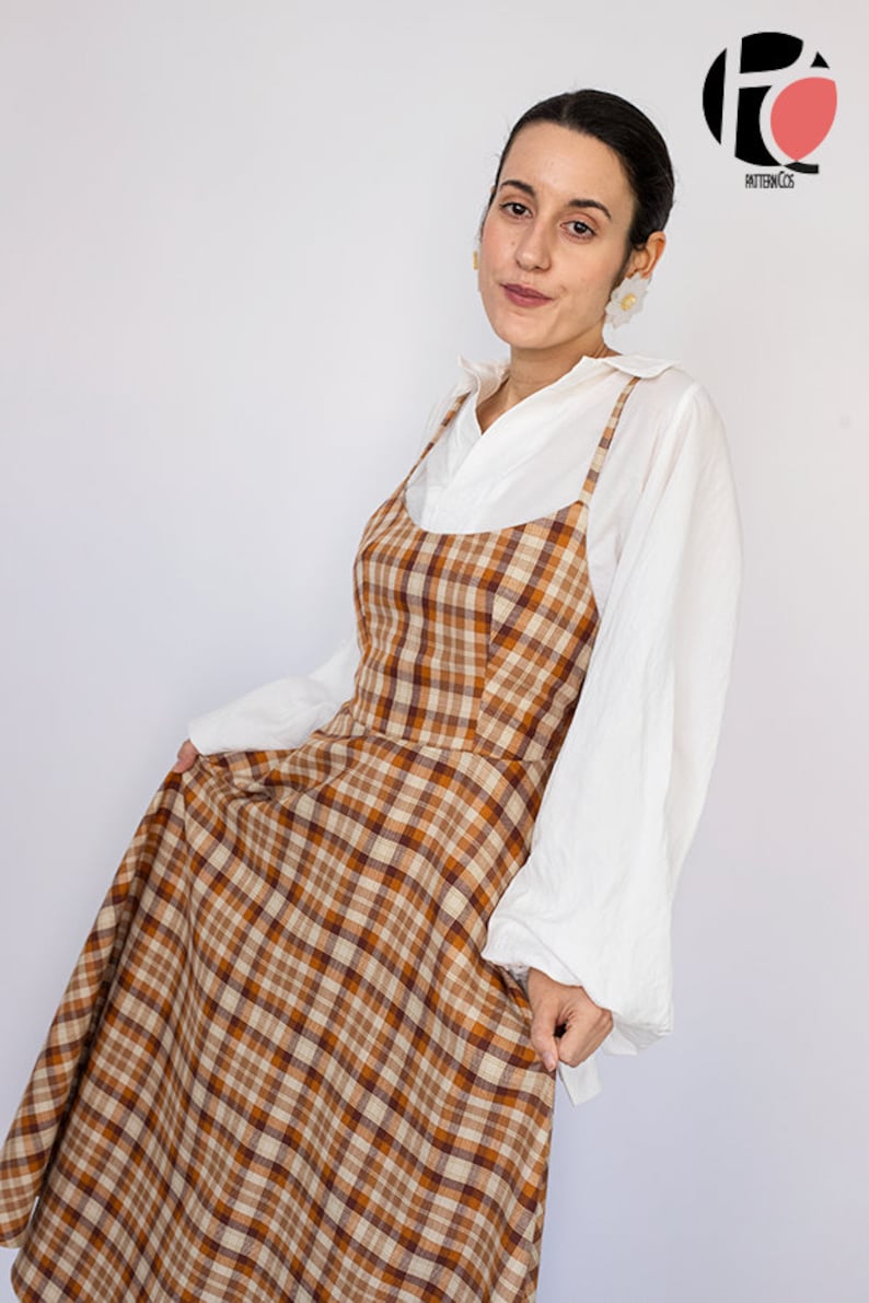 Pode incluir: Uma mulher vestindo um vestido de avental xadrez marrom e branco sobre uma camisa branca abotoada. O vestido tem um decote redondo e al&ccedil;as finas. A mulher est&aacute; segurando a saia do vestido com a m&atilde;o direita.