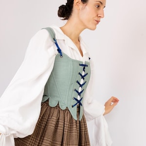 May include: A light green corset with blue lacing and straps is worn over a white blouse and a brown plaid skirt. The corset has a scalloped edge. The outfit is reminiscent of historical fashion.