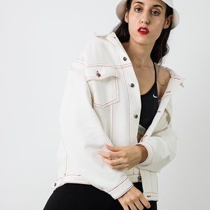 Pu&ograve; includere: Una giacca di jeans bianca con cuciture rosse, indossata da una persona con un cappello a secchiello bianco e leggings neri. La giacca ha una chiusura con bottoni e una tasca sul lato sinistro.