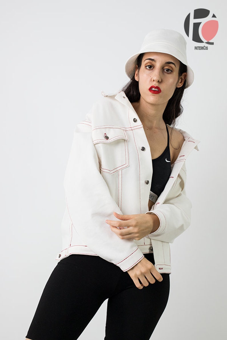 Pu&ograve; includere: Una giacca di jeans bianca con cuciture rosse, indossata da una persona con un cappello a secchiello bianco e leggings neri. La giacca ha una chiusura con bottoni e una tasca sul lato sinistro.