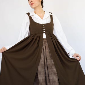 May include: A woman wearing a brown dress with a white collared shirt underneath. The dress has a square neckline and button details. The skirt is a brown plaid pattern. The dress is a vintage style.