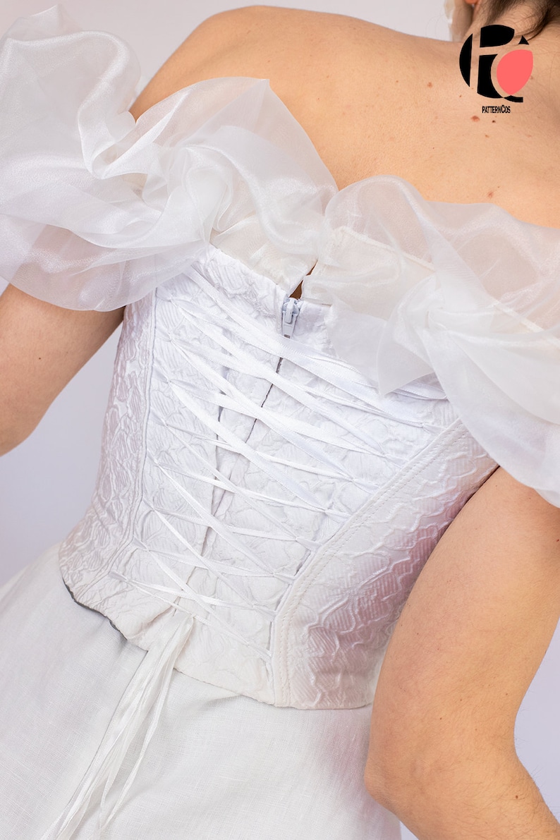 May include: White corset with a lace-up back and a ruffled off-the-shoulder neckline. The corset is made of a textured fabric and has a zipper closure in the back.