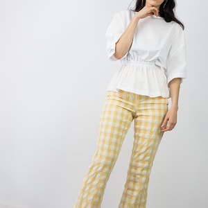 May include: A woman wearing a white, loose-fitting blouse with a gathered waist and yellow and white gingham pants. She is wearing black heeled sandals.