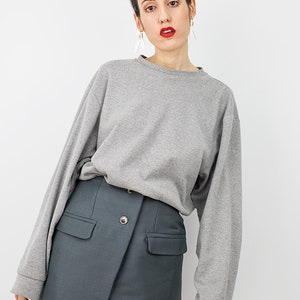 May include: A woman wearing a gray long-sleeved sweatshirt and a dark gray skirt with a button closure. The skirt has two pockets on the front.