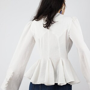 May include: White long-sleeved blouse with a ruffled peplum hem and gathered sleeves. The blouse has a fitted bodice and a slightly flared skirt.