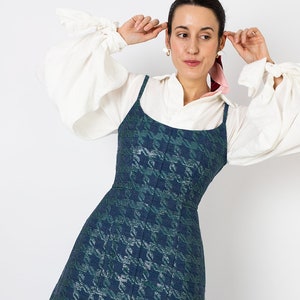 K&ouml;nnte beinhalten: Ein &auml;rmelloses, dunkelblau-gr&uuml;nes Kleid mit Hahnentrittmuster und Spaghettitr&auml;gern. Das Kleid wird &uuml;ber einer wei&szlig;en Bluse mit weiten &Auml;rmeln getragen. Das Model tr&auml;gt goldene Ohrringe und einen rosa Schal.