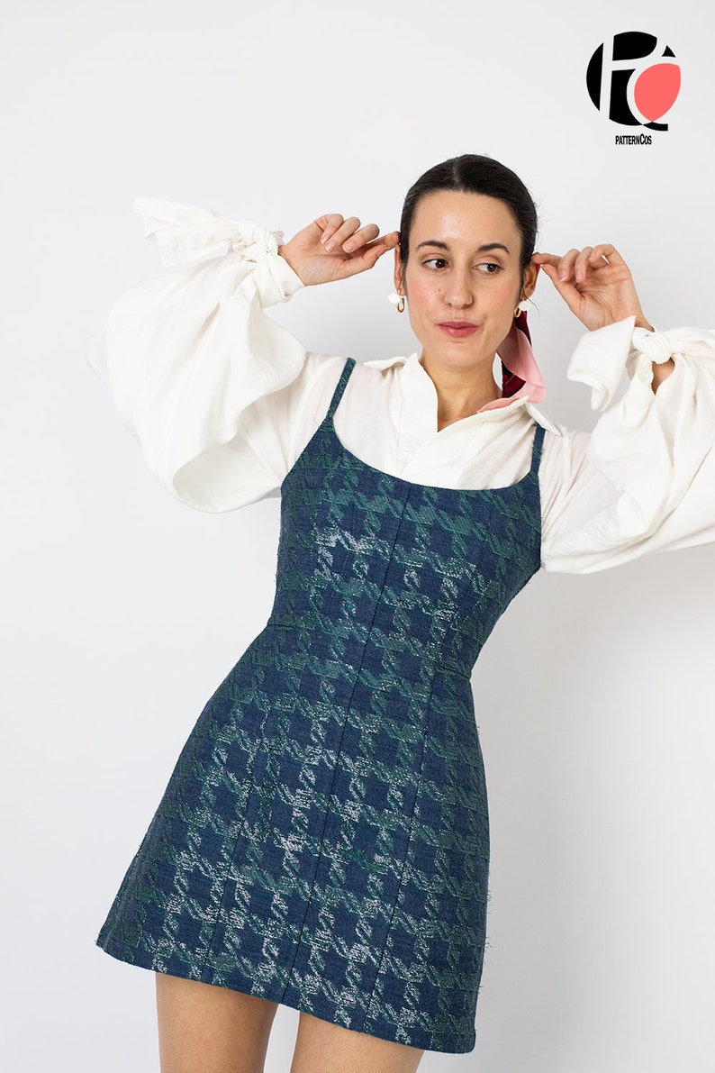 K&ouml;nnte beinhalten: Ein &auml;rmelloses, dunkelblau-gr&uuml;nes Kleid mit Hahnentrittmuster und Spaghettitr&auml;gern. Das Kleid wird &uuml;ber einer wei&szlig;en Bluse mit weiten &Auml;rmeln getragen. Das Model tr&auml;gt goldene Ohrringe und einen rosa Schal.