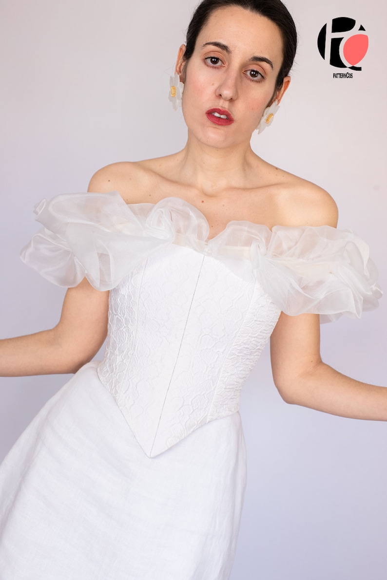 May include: A white off-the-shoulder corset with a ruffled neckline and a lace pattern. The corset is paired with a white skirt.