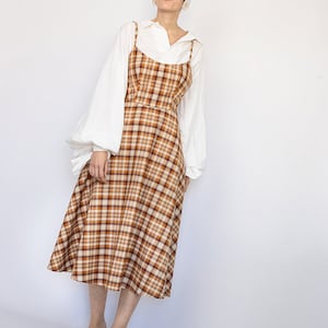 Pode incluir: Uma mulher vestindo uma blusa branca com um vestido midi xadrez marrom e bege e sand&aacute;lias verdes. O vestido tem al&ccedil;as finas e um corpete ajustado.