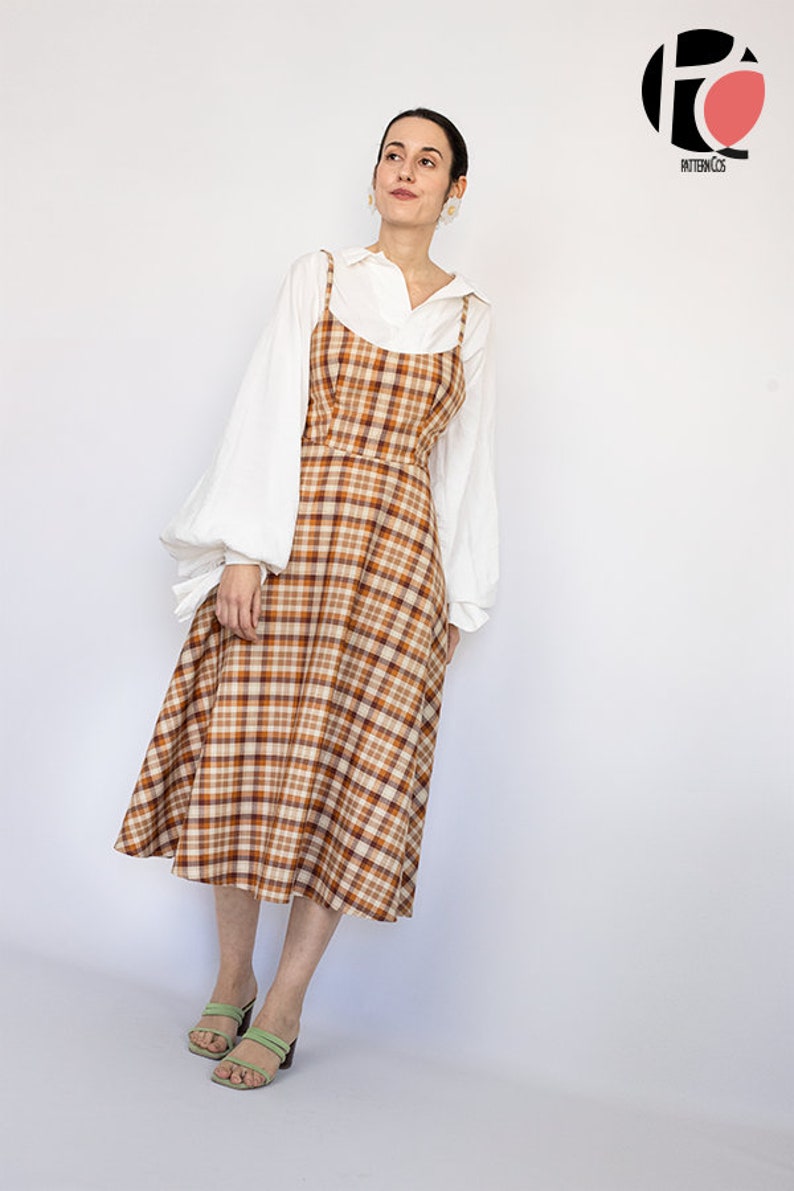 Pode incluir: Uma mulher vestindo uma blusa branca com um vestido midi xadrez marrom e bege e sand&aacute;lias verdes. O vestido tem al&ccedil;as finas e um corpete ajustado.