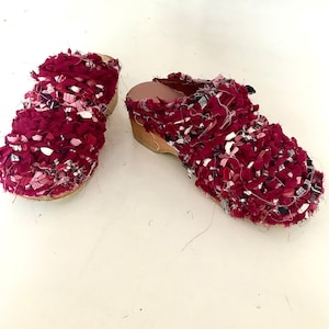 Op de afbeelding: Een paar bordeauxrode klompen met een bovenwerk van stof. De stof is een mix van bordeauxrode, witte en roze stroken. De klompen hebben een houten zool en een kleine hak. De schoenen staan op een witte ondergrond.