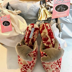 Puede incluir: Un par de zapatillas de terciopelo con estampado floral rojo y dorado y detalle de lazo. Los zapatos están dentro de una caja con papel de seda y una etiqueta rosa con el texto "Leigh Morrison Footwear".