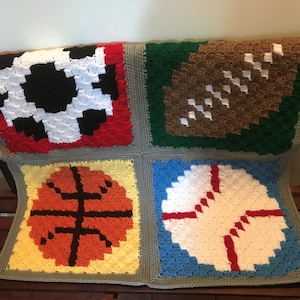 May include: A crocheted blanket with four squares, each featuring a different sport: soccer, football, basketball, and baseball. The squares are in red, green, yellow, and blue, respectively.