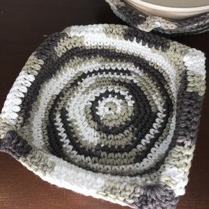 May include: Handmade crochet bowl cozy in shades of white, gray, and olive green. The cozy has a square shape with rounded corners and a spiral pattern. The second bowl cozy is visible in the background.